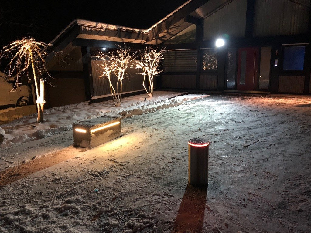 Versenkbarer Edelstahlpoller mit rotem LED-Leuchtring in verschneiter Einfahrt vor modernem Haus bei Nacht – offene Architektur ohne Zaun