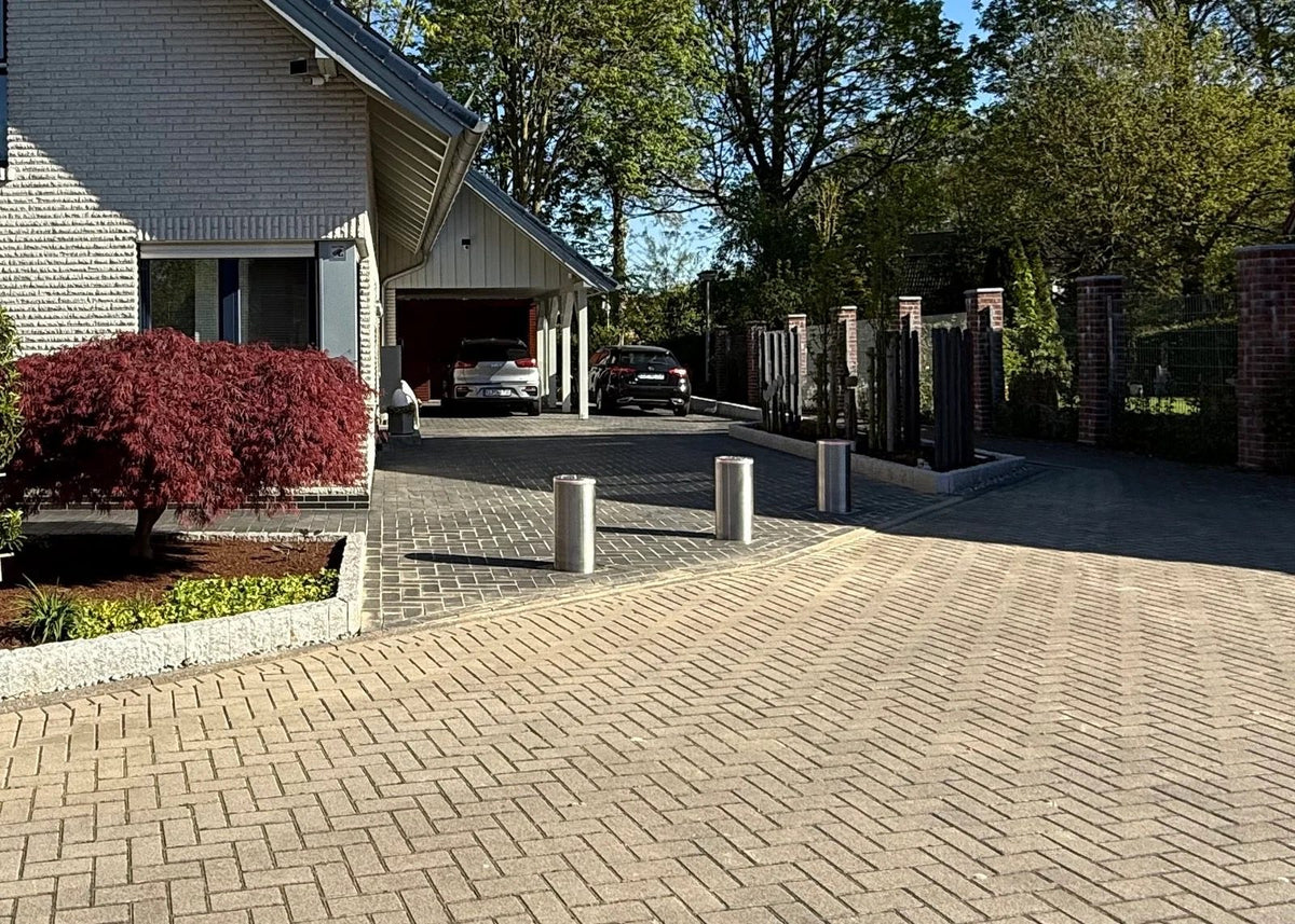 Drei ausgefahrene versenkbare Edelstahl-Poller sperren die breite gepflasterte Einfahrt eines Wohnhauses vor Carport und Garage