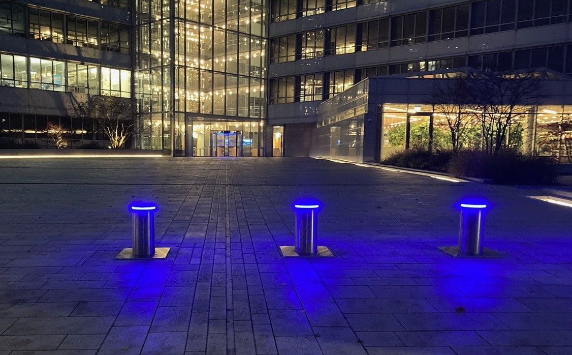 Drei ausgefahrene versenkbare Sicherheitspoller aus Edelstahl mit blauem LED-Leuchtring sichern nachts den Vorplatz der Firmenzentrale eines Finanzdienstleisters