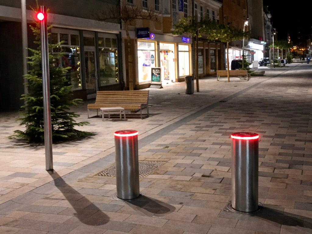 Zwei ausgefahrene versenkbare Sicherheitspoller aus Edelstahl V4A mit roter LED-Beleuchtung an der Zufahrt zu einer Fußgängerzone bei Nacht, daneben eine Ampel auf Rot