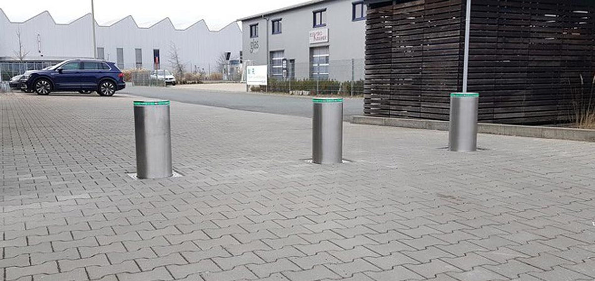 Drei ausgefahrene versenkbare Edelstahl-Poller mit grünem LED-Ring sichern die gepflasterte Zufahrt eines Firmengeländes im Gewerbegebiet