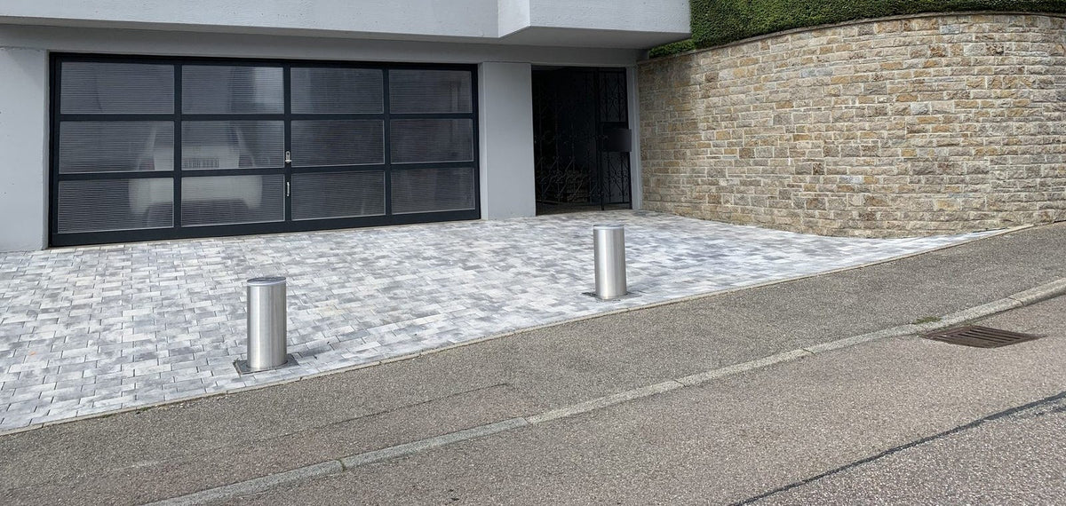 Zwei ausgefahrene versenkbare Poller aus Edelstahl V4A am Straßenrand der gepflasterten Einfahrt eines modernen Hanghauses mit Glas-Garagentor und Natursteinmauer