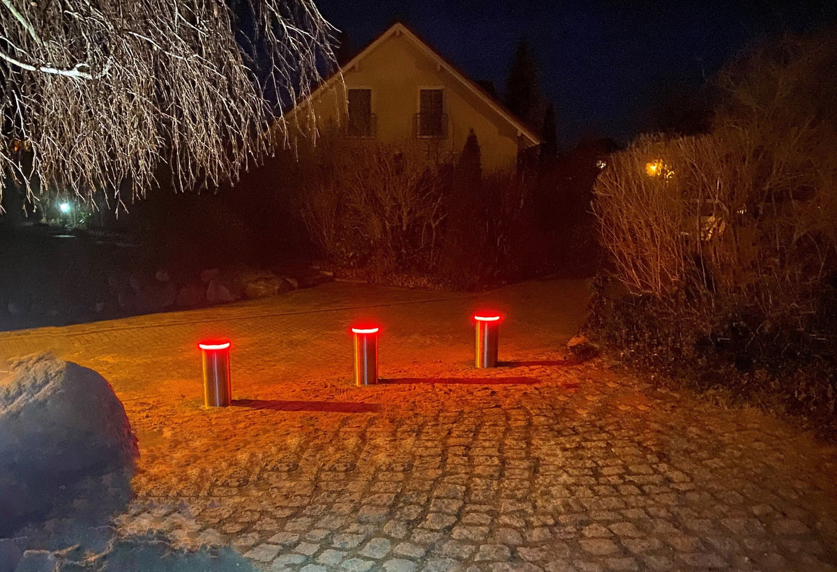 Drei ausgefahrene versenkbare Edelstahl-Poller mit rot leuchtendem LED-Ring sperren nachts eine breite Einfahrt aus Granitpflaster vor einem Wohnhaus