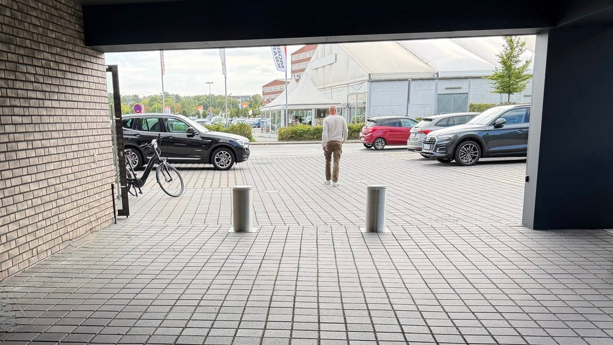 Zwei ausgefahrene Edelstahl-Poller in der Durchfahrt einer Wohnanlage, dahinter der Parkplatz eines Supermarkts