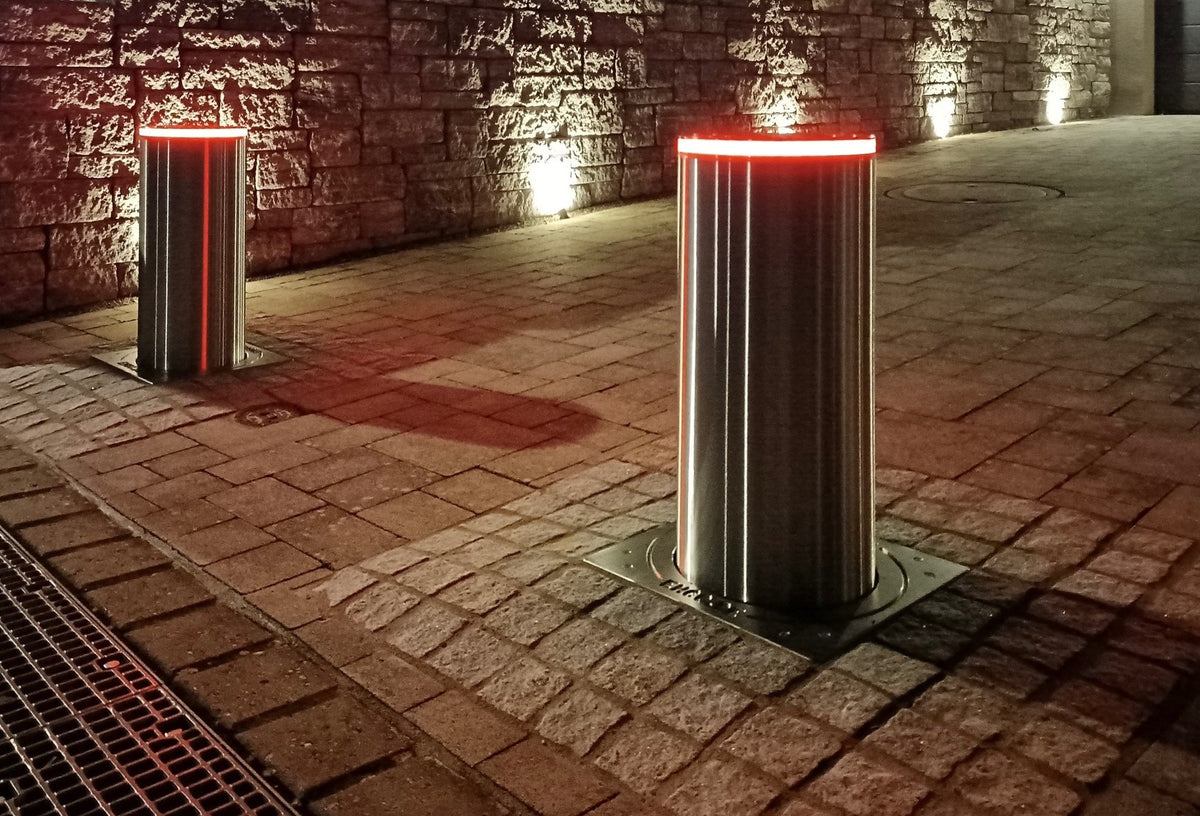 Zwei versenkbare Edelstahl-Poller mit rot leuchtendem LED-Ring sichern nachts eine gepflasterte Einfahrt vor einer beleuchteten Natursteinmauer