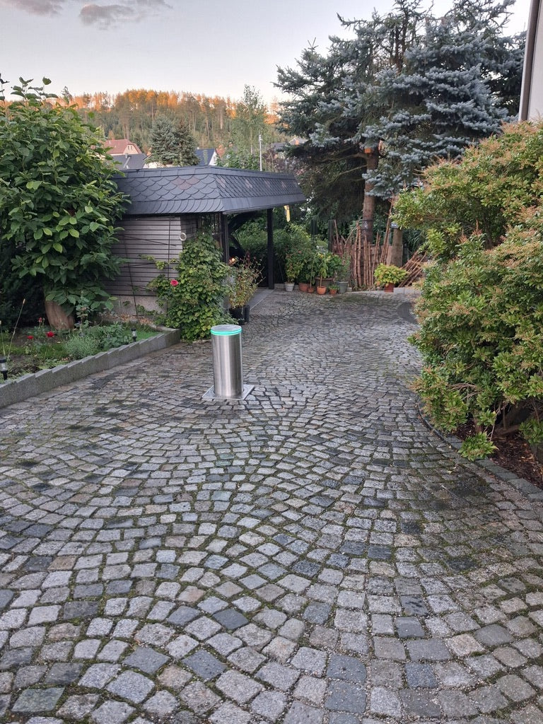 Ausgefahrener versenkbarer Poller aus Edelstahl V4A mit grünem LED-Ring in einer Einfahrt aus Granit-Kleinpflaster vor einem Carport