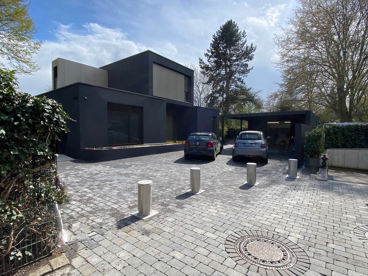 Vier ausgefahrene versenkbare Edelstahl-Poller sichern die gepflasterte Einfahrt eines modernen Eigenheims mit Carport – Schutz vor Fremdparkern und Autodiebstahl