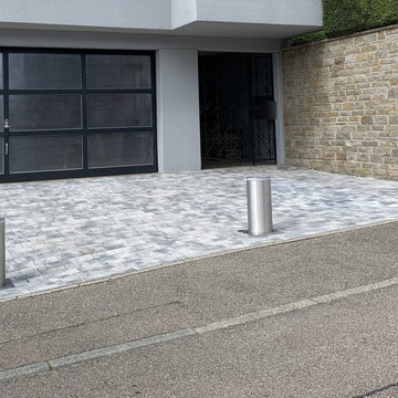Zwei ausgefahrene versenkbare Poller aus Edelstahl V4A am Straßenrand der gepflasterten Einfahrt eines modernen Hanghauses mit Glas-Garagentor und Natursteinmauer