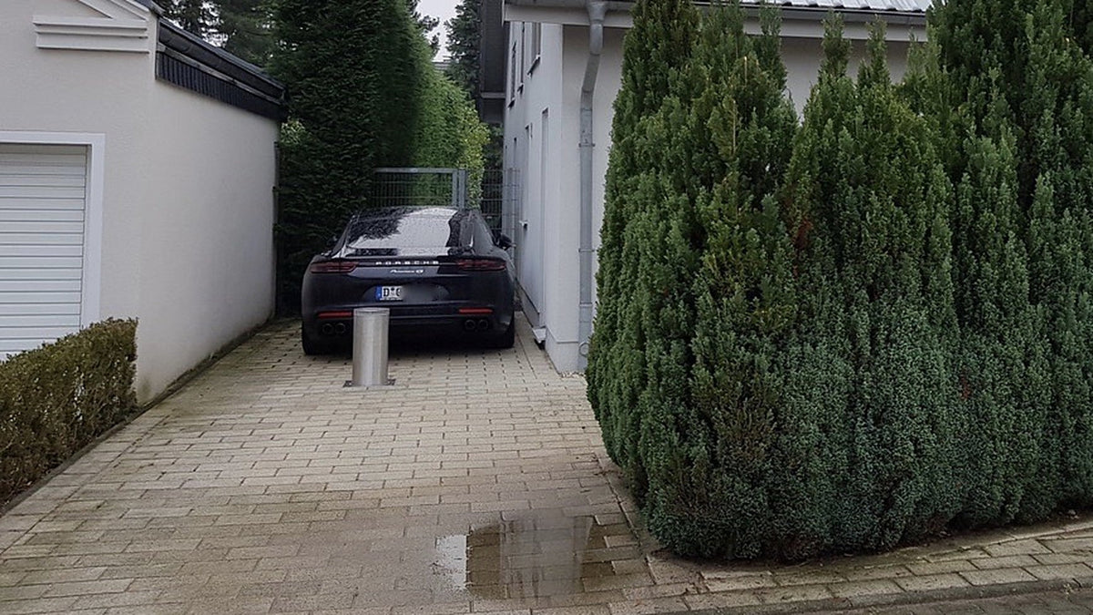 Versenkbarer Edelstahl-Poller sperrt eine schmale gepflasterte Einfahrt zwischen Hecke und Garage, dahinter ein geparkter Sportwagen